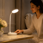 Femme lisant à un bureau clair, lampe à col flexible allumée près d'un bouquet blanc.