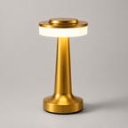 Lampe dorée allumée vue de trois quarts sur fond gris, base évasée et pied fin.