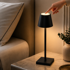 Main effleurant le haut de la lampe noire allumée posée près d'un lit aux draps clairs.