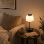 Lampe allumée sur un guéridon rond dans un salon beige, près d'un fauteuil et de branchages.
