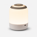 Lampe cylindrique allumée vue de trois quarts, base beige et prise de recharge latérale.